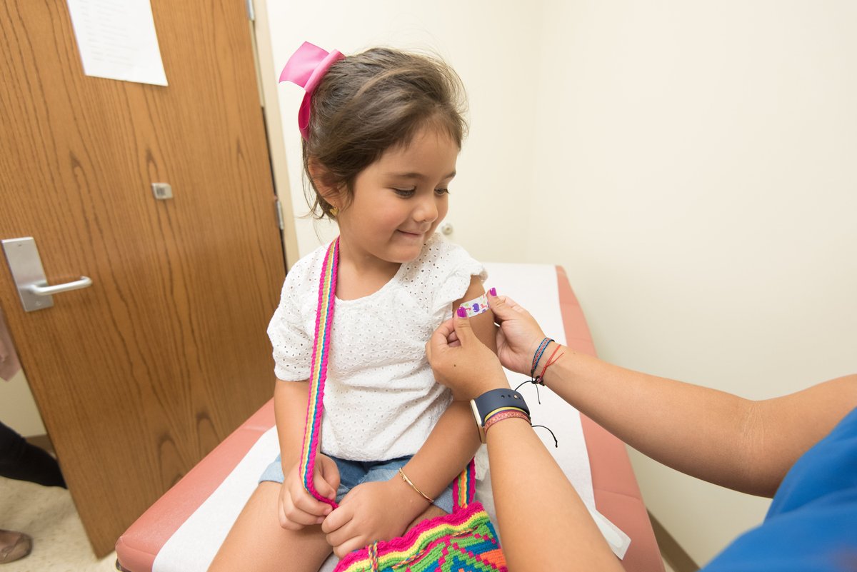 A smiling young girl receives an adhesive bandage on her arm from a health worker in an exam room.