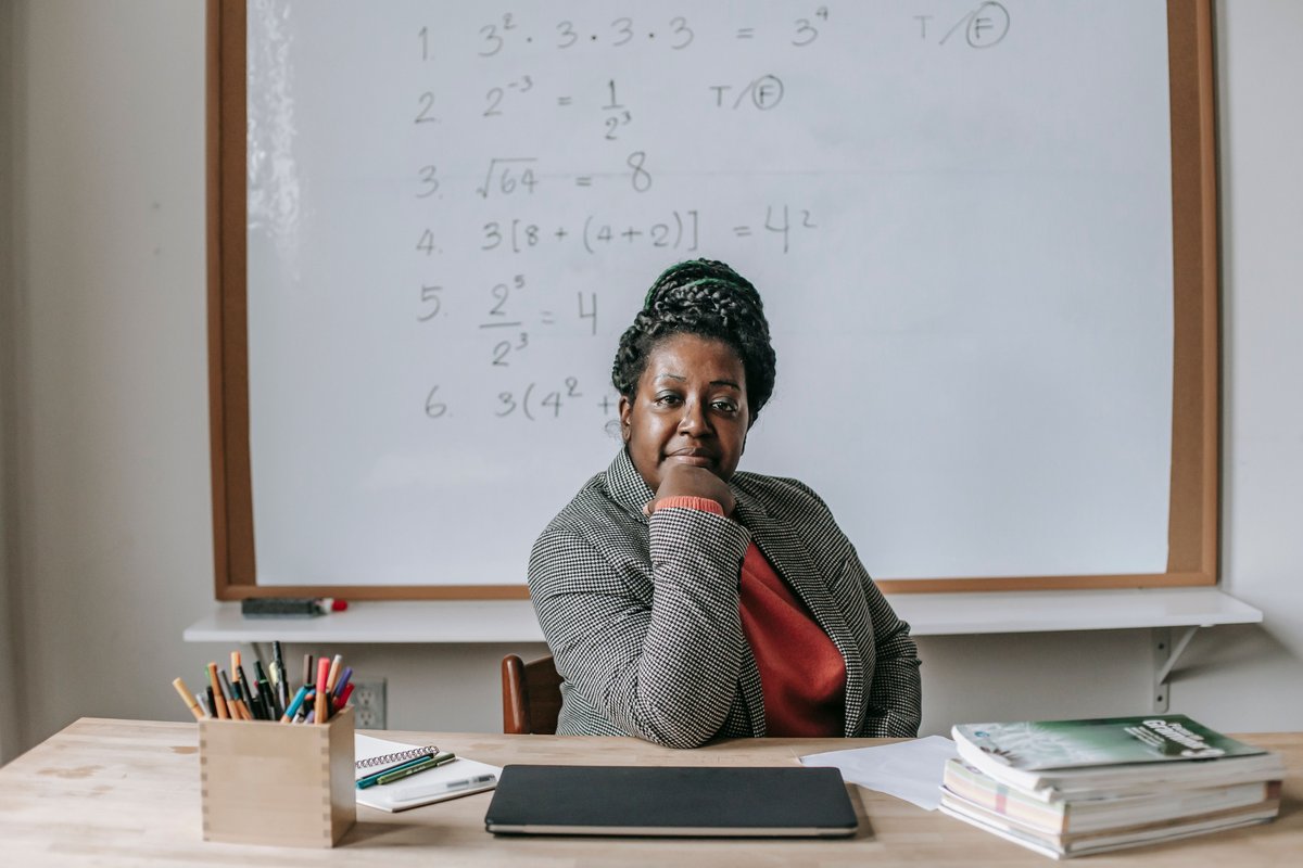 A teacher seated at a desk in front of a whiteboard with math problems.